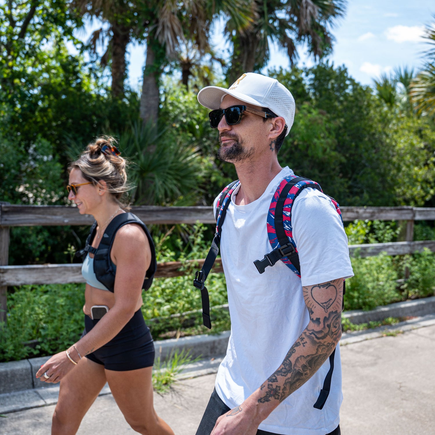 Two people wearing Ruck Plate Carrier 3.0 backpacks with ergonomic lumbar support stroll outdoors on a sunny day, surrounded by greenery and palm trees.