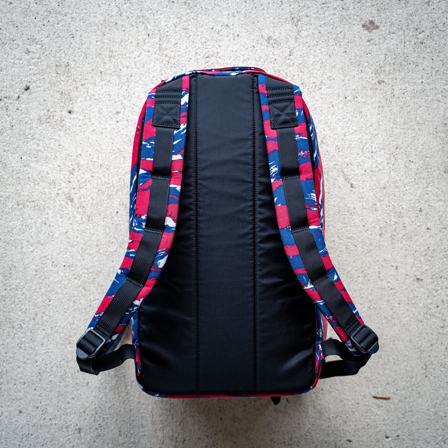 The Bullet Ruck Classic - Ballistic Nylon Cordura is a streamlined 15L daypack with blue, red, and white patterned straps and back, shown laid flat on a light concrete surface.