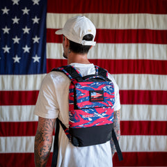 A man wearing a Ruck Plate Carrier 3.0 featuring ergonomic lumbar support stands in front of a large American flag.