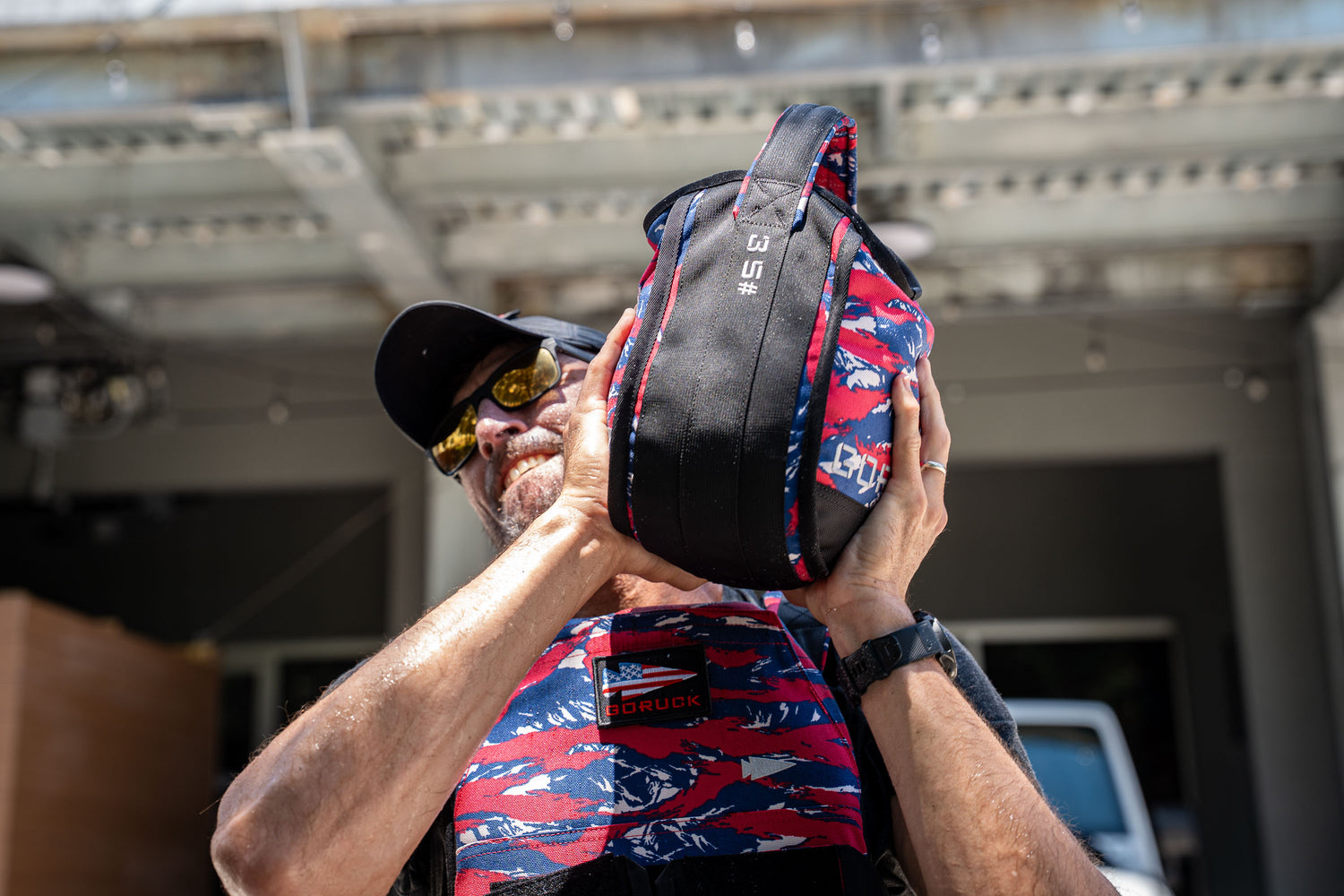 A man in sunglasses lifts a colorful Sand Kettlebells outdoors while wearing a weighted vest and cap. The 35 lb portable kettlebell features a padded handle for comfort.