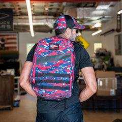 A man in a workshop, facing away from the camera, wears a red, white, and blue camo Rucker 5.0 rucksack with a matching cap.