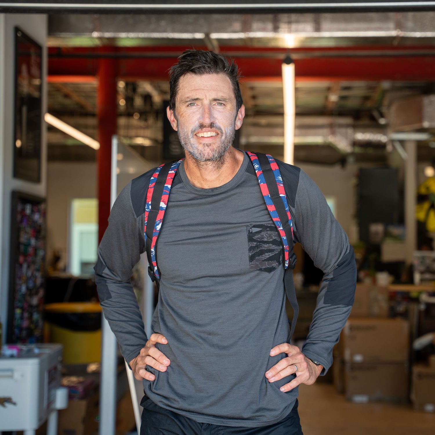 A man with a beard, in a gray long-sleeve shirt, stands indoors with hands on hips wearing the Bullet Ruck Classic - Ballistic Nylon Cordura 15L backpack.