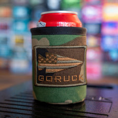A red can in a camo Ballistic Beer Jacket with a spear-and-stars patch sits against a blurred colorful background—a rugged, customizable sleeve for your drink on any adventure.