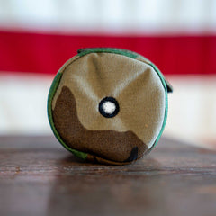 A small, round camouflage Ballistic Beer Jacket—customizable beer sleeve with a black-bordered white circle on the front—rests on a wooden surface.