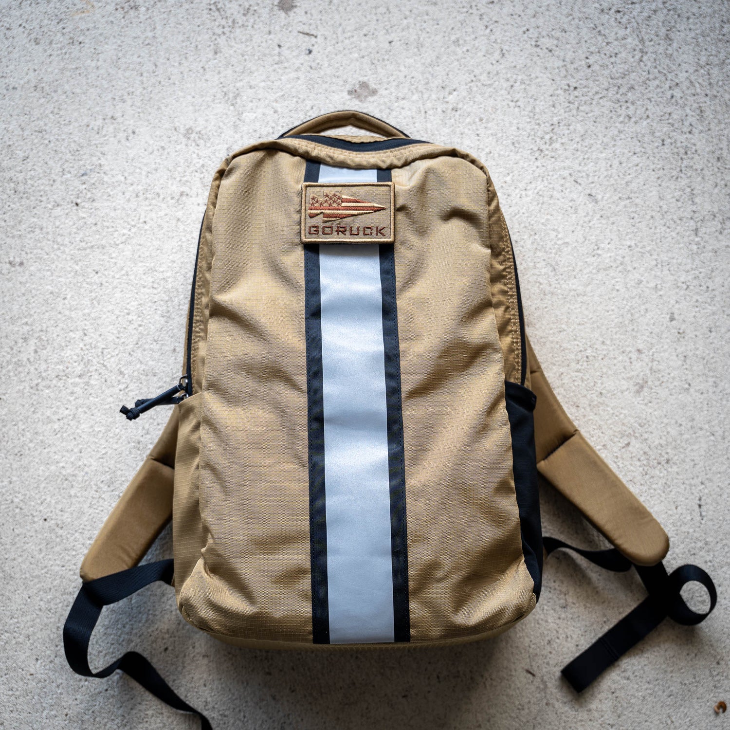 The Basic Rucker backpack, featuring a tan design with a reflective gray stripe, a GORUCK patch, and space for a ruck plate, rests on a light concrete surface—ideal for your next rucking adventure.