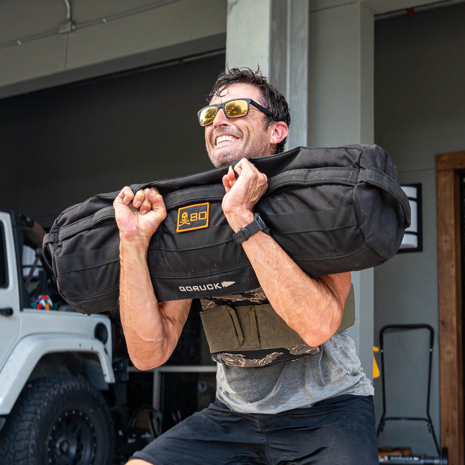 Wearing sunglasses and lifting a weighted sandbag outdoors, this man is all smiles as he enjoys his rucking workout—made even more intense with the Rucking Weight Vest.