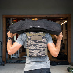Person lifting a black GORUCK sandbag onto their shoulders, wearing the Rucking Weight Vest in a camo pattern.