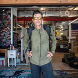 A man in sunglasses and a green Warm Up Full Zip jacket with ToughDry shoulders stands inside a room decorated with flags and gear, his lightweight jacket and backpack hinting at adventure-ready style.