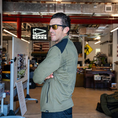 A man wearing sunglasses and the Warm Up Full Zip jacket in green stands indoors with his arms crossed, looking to the side. The lightweight jacket features zippered pockets and ToughDry shoulders for added durability.