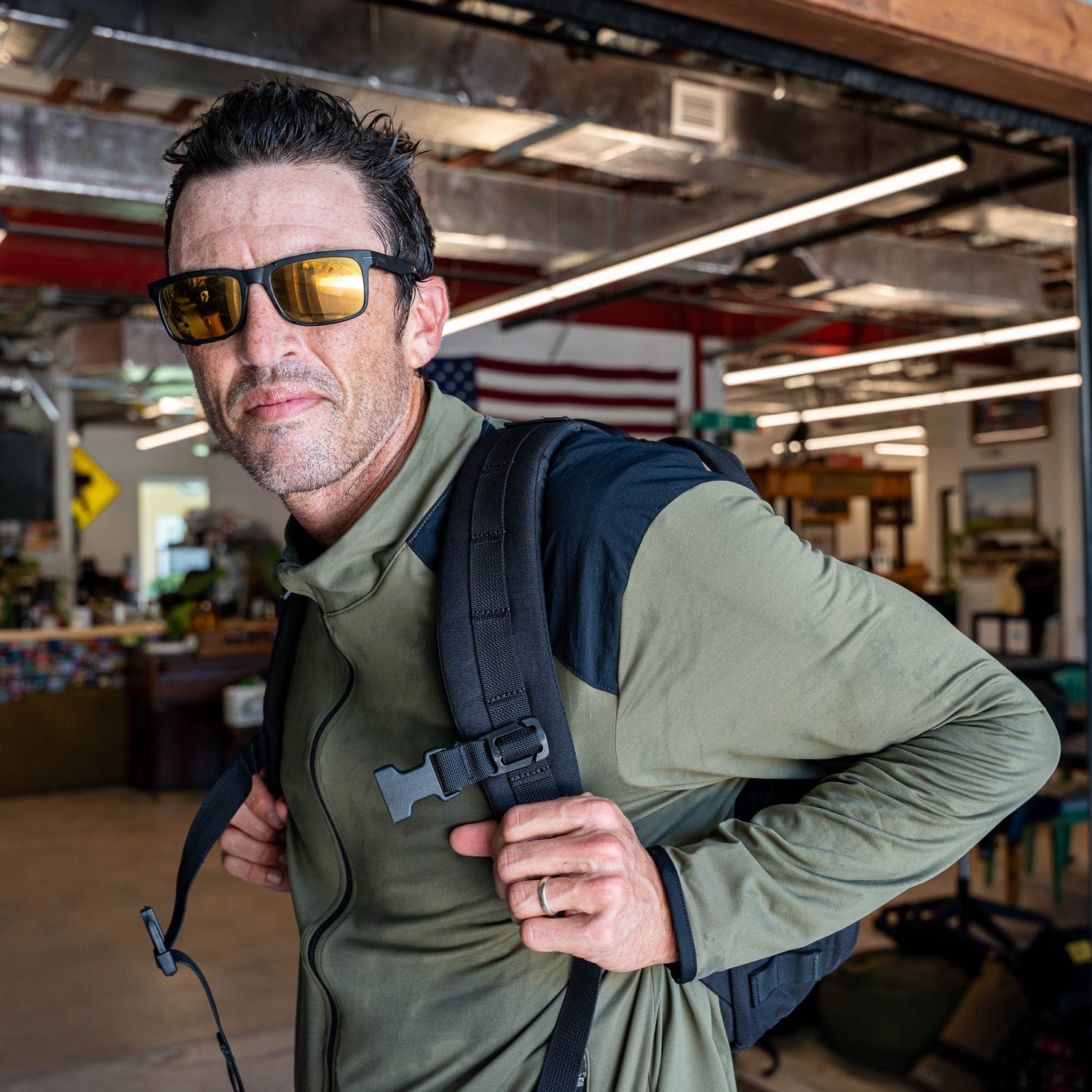 A man wearing sunglasses and the Warm Up Full Zip—a green, lightweight jacket with ToughDry shoulders—stands indoors with a backpack, an American flag visible in the background. The Warm Up Full Zip is proudly made in the USA.