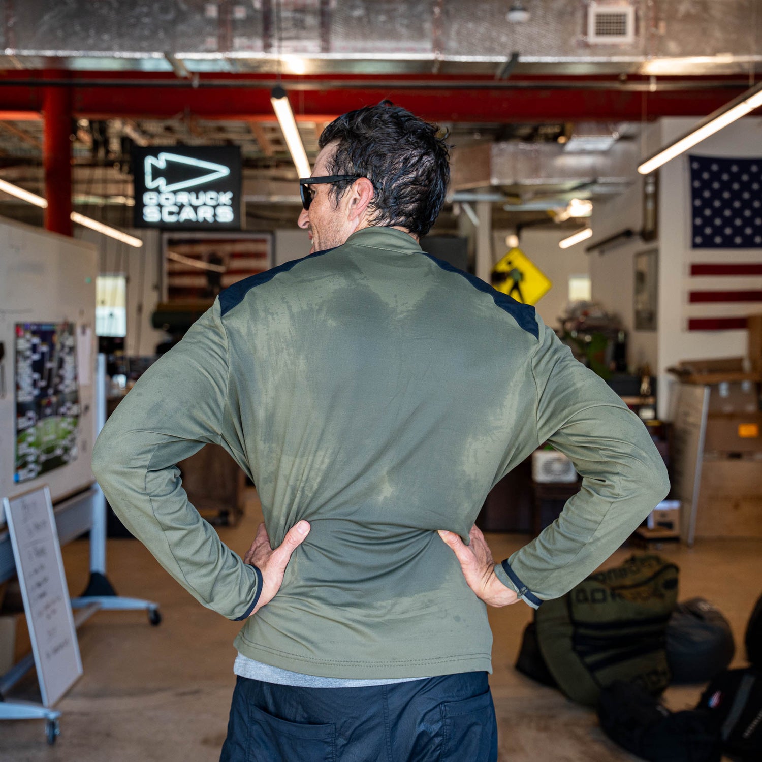 A man wearing the Warm Up Full Zip olive lightweight jacket with zippered pockets stands with his hands on his hips in an indoor workspace featuring an American flag.