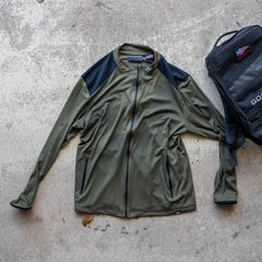 Warm Up Full Zip in olive green with ToughDry shoulders and black shoulder panels, shown next to a black backpack on a concrete surface.