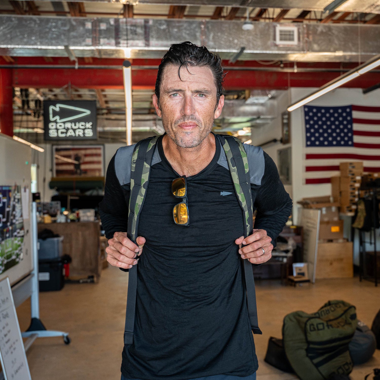 A man stands indoors with an American flag behind him, wearing sunglasses and a black shirt, carrying the Bullet Ruck Classic - Ballistic Nylon Cordura packable backpack.