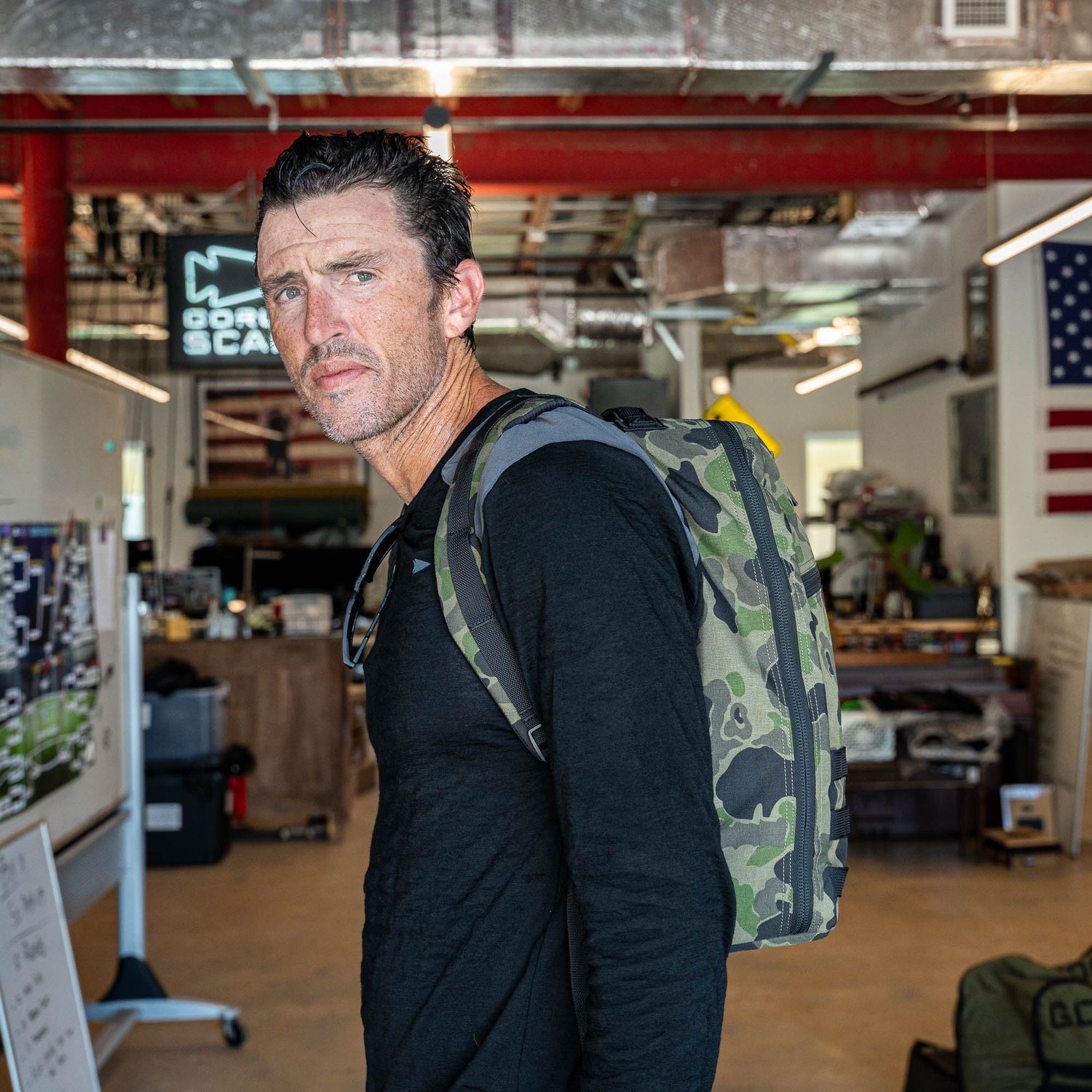 A man wearing a black shirt with the Bullet Ruck Classic - Ballistic Nylon Cordura 15L daypack stands in an industrial-style room with flags and whiteboards.