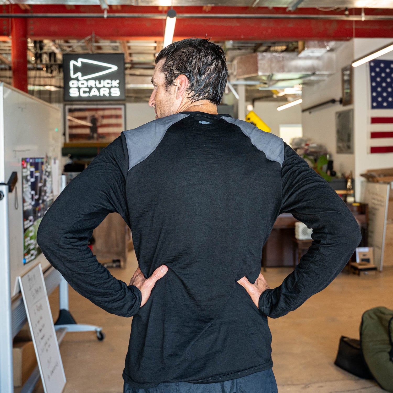 A man stands indoors with his hands on his hips, facing away, wearing the Men’s Commando Long Sleeve - Merino Wool shirt in black with gray reinforced shoulders reminiscent of a classic commando sweater.
