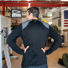 A man stands indoors with his hands on his hips, facing away, wearing the Men’s Commando Long Sleeve - Merino Wool shirt in black with gray reinforced shoulders reminiscent of a classic commando sweater.