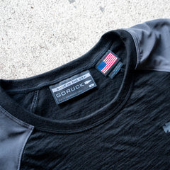 Men’s Commando Long Sleeve - Merino Wool in black and gray, featuring a USA flag tag and "Built in the USA" label, displayed on a concrete surface. Designed with reinforced shoulders inspired by the classic commando sweater for enhanced durability.