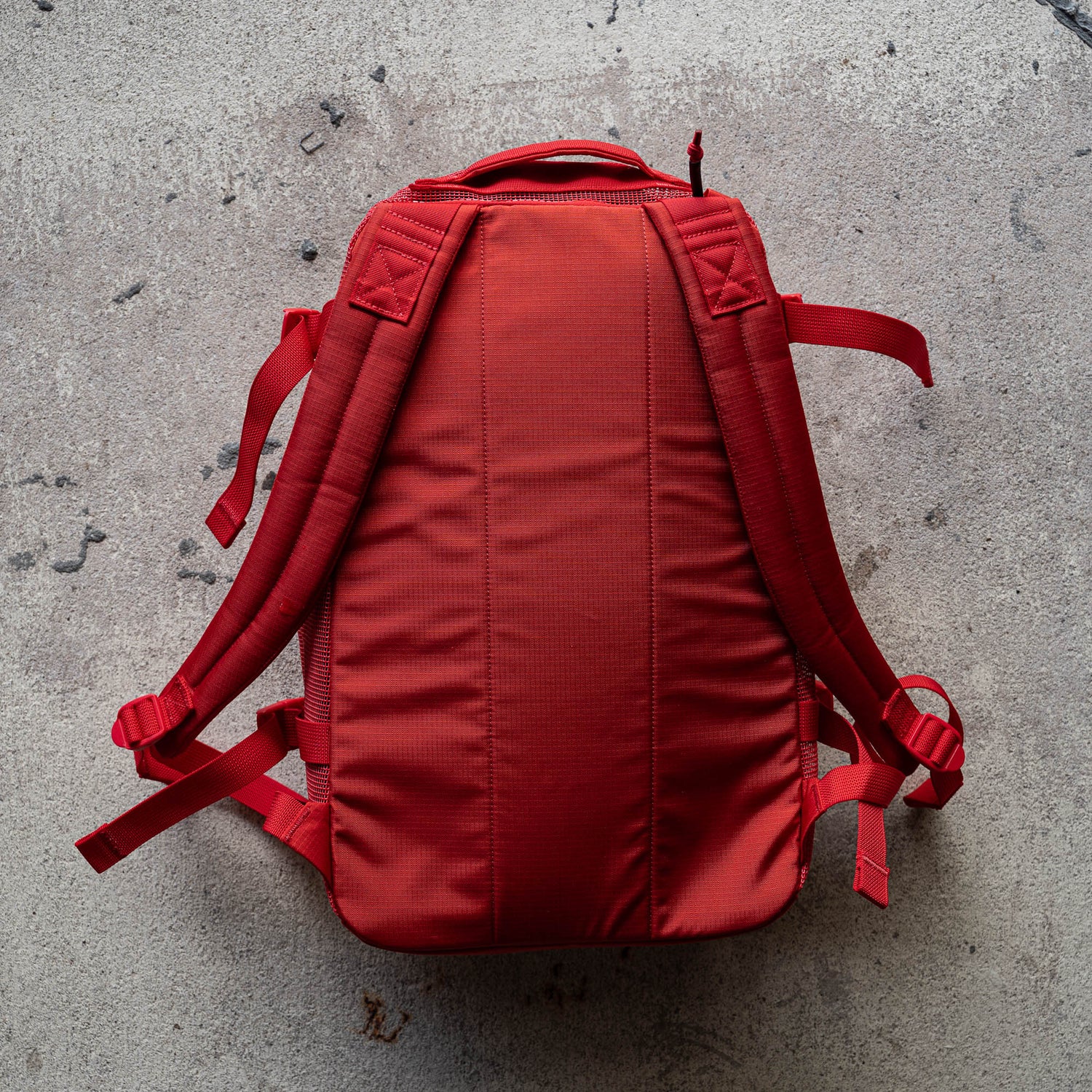 A red Mesh Ruck with padded straps and quick-drying material is lying flat on a textured concrete floor.