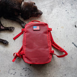 A red Mesh Ruck backpack with a USA flag patch lies on concrete next to a resting black dog.