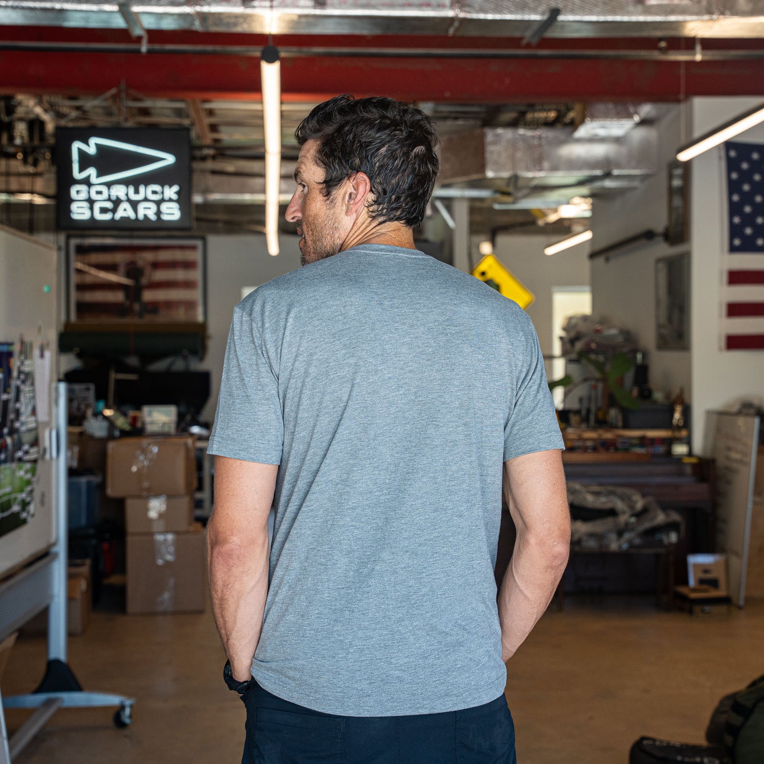 A man wearing the Slick Short Sleeve Tee - Tri-BlendX in gray stands in a workshop, facing away; an American flag and 
