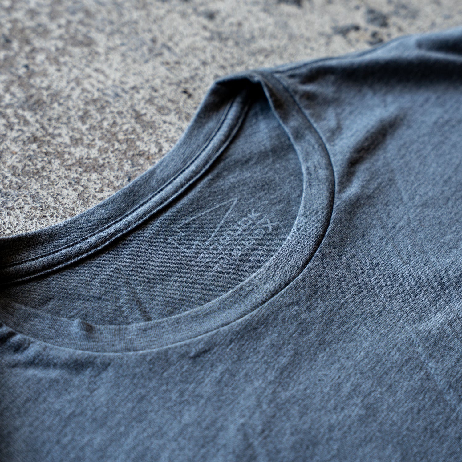 Close-up of the collar on the Slick Short Sleeve Tee - Tri-BlendX in gray, highlighting the inner neck label against a rough textured background.