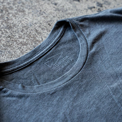 Close-up of the collar on the Slick Short Sleeve Tee - Tri-BlendX in gray, highlighting the inner neck label against a rough textured background.