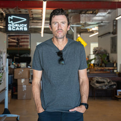 A man wearing the Slick Short Sleeve Tee - Tri-BlendX in gray stands in an industrial office, with boxes and a GORUCK SCARS sign behind him.