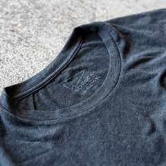 Close-up of a black Slick Short Sleeve Tee - Tri-BlendX collar with the "GORUCK" brand label visible, lying on a textured surface. The premium Tri-BlendX fabric features an antimicrobial treatment for lasting freshness.