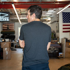 A man wearing a dark Slick Short Sleeve Tee - Tri-BlendX stands with arms crossed in a cluttered room, facing away from the camera.