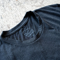 Close-up of the black Slick Short Sleeve Tee - Tri-BlendX collar with a printed label displaying the "GORUCK" logo and sizing details on concrete, showcasing its premium Tri-BlendX material for superior comfort.