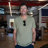A man stands smiling in a garage with his hands in his pockets and sunglasses on his shirt, wearing the tan Slick Short Sleeve Tee - Tri-BlendX.