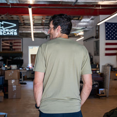 A man wearing the Slick Short Sleeve Tee - Tri-BlendX in olive stands in a cluttered room decorated with American flags, facing away.