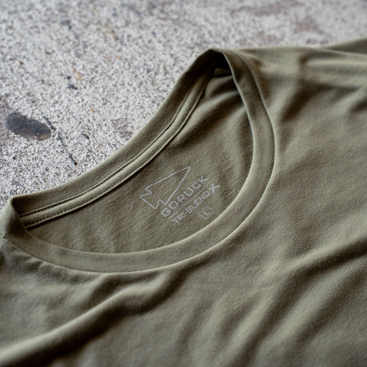 A close-up of an olive green Slick Short Sleeve Tee - Tri-BlendX featuring a GORUCK logo and label on the inside collar, lying on concrete.