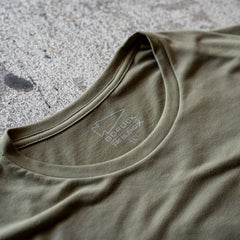A close-up of an olive green Slick Short Sleeve Tee - Tri-BlendX featuring a GORUCK logo and label on the inside collar, lying on concrete.
