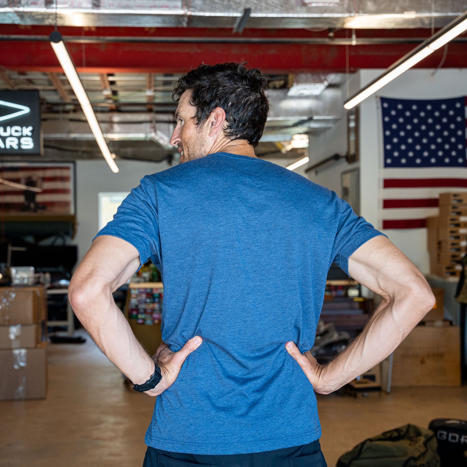 A man wearing the Slick Short Sleeve Tee - Tri-BlendX in blue stands with his hands on his hips in a warehouse adorned with American flags.