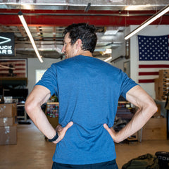 A man wearing the Slick Short Sleeve Tee - Tri-BlendX in blue stands with his hands on his hips in a warehouse adorned with American flags.