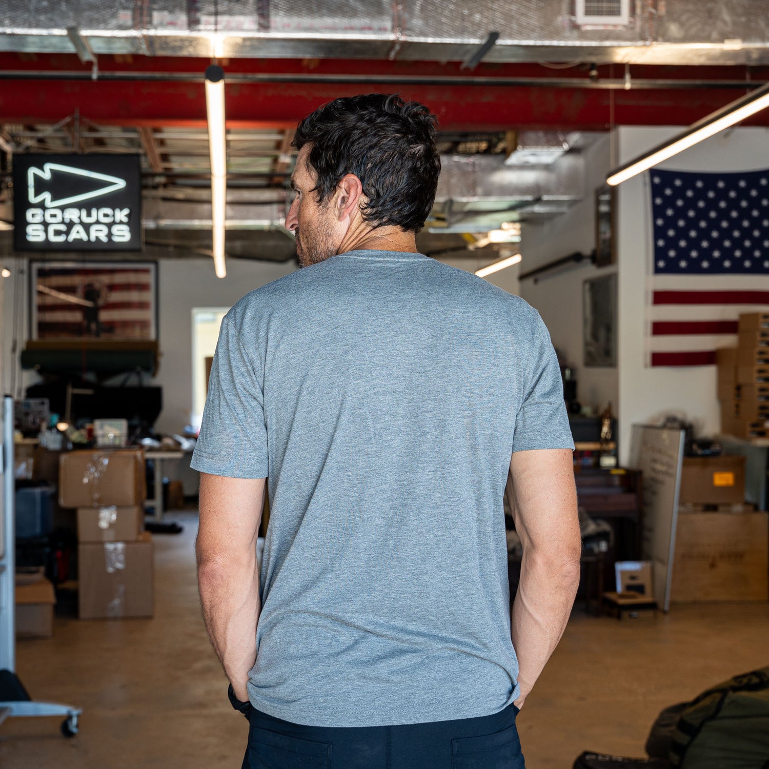 Wearing the Short Sleeve Pocket Tee - Tri-BlendX, a man stands with his back to the camera in a workshop adorned with an American flag, embodying classic simplicity and that versatile, 