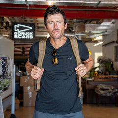 A man wearing a travel backpack and sunglasses on his shirt stands in a workshop with a "GORUCK SCARS" sign in the background, showcasing the Double Bullet Ruck w/ Bombproof Laptop Compartment - 18L.