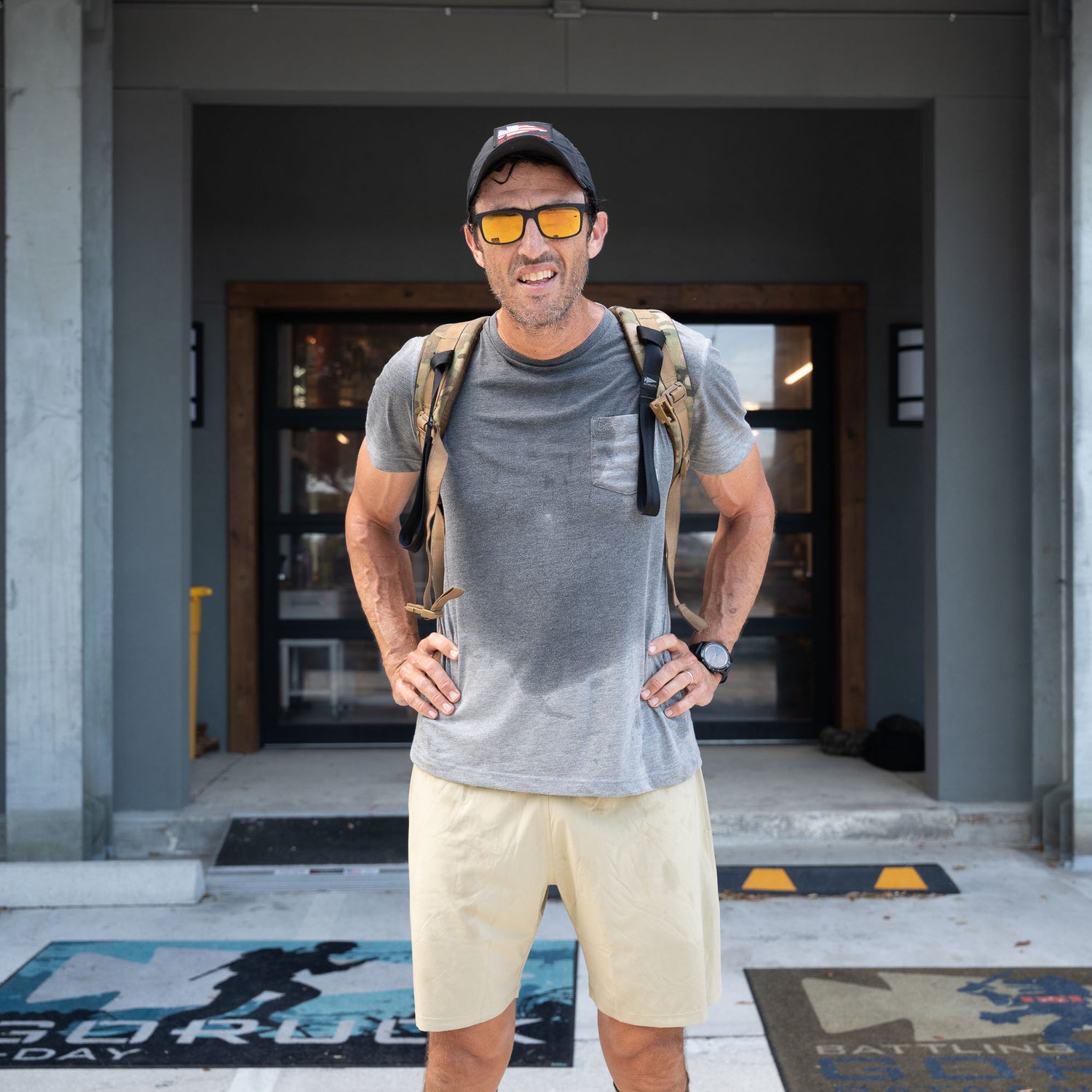 A man in sunglasses, cap, and backpack stands outside a building, his Men’s Training Shorts - ToughStretch—with a built-in liner for added comfort—paired with a shirt visibly soaked with sweat.