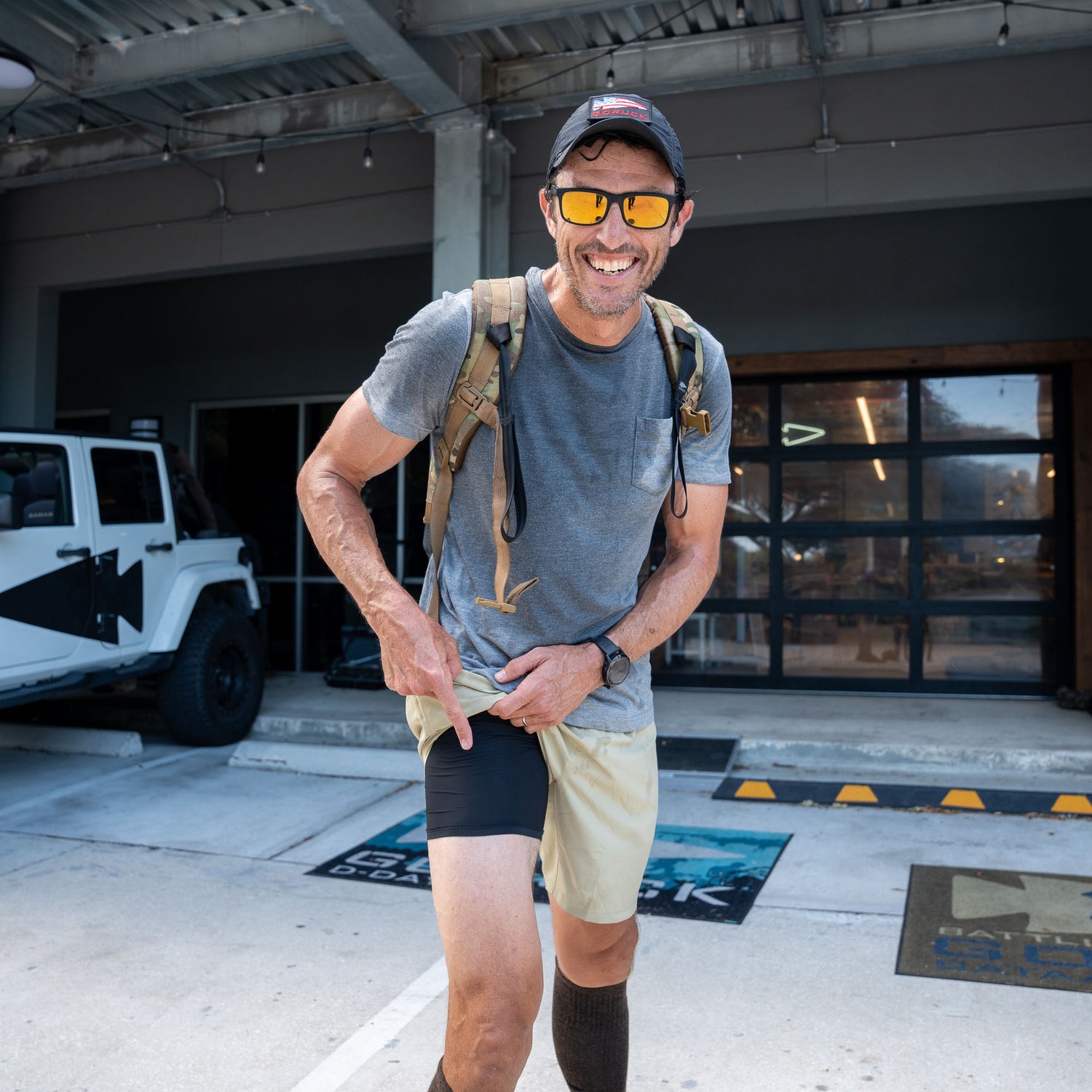 A smiling man in sunglasses and a cap stands outside near a white Jeep, pointing to his black Men’s Training Shorts - ToughStretch.