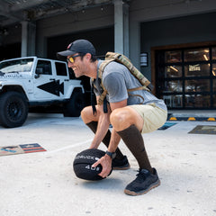 A man in athletic gear squats outdoors, lifting a 40 lb medicine ball near a white Jeep, wearing Men’s Training Shorts - ToughStretch with a built-in liner for added comfort and support.