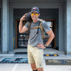 A man in athletic wear and sunglasses smiles, holding a sandbag over his shoulder outside a building, confidently showcasing his Men’s Training Shorts - ToughStretch with a supportive built-in liner.