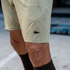Close-up of a person wearing Men’s Training Shorts - ToughStretch in light tan with a built-in liner and dark brown socks, standing on a concrete surface.
