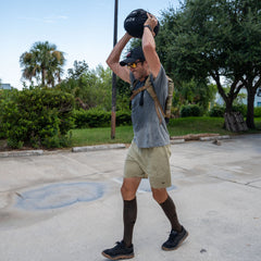 A man outdoors lifts a weighted sandbag overhead, wearing a backpack, hat, and Men’s Training Shorts - ToughStretch with a built-in liner on a sunny day.