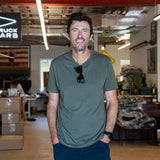 A man in a green Slick Short Sleeve Tee - Tri-BlendX and sunglasses stands smiling in a workshop with boxes and tools in the background.
