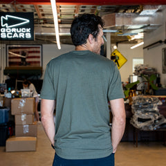 A man wearing the Slick Short Sleeve Tee - Tri-BlendX in olive stands with his back to the camera in a workspace featuring boxes and a GORUCK sign, highlighting this durable, antimicrobial shirt designed for active environments.