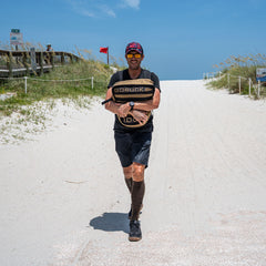 A man performs loaded carries with a Sand Tombstone along a sandy beach path beneath a clear blue sky.