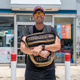 A smiling man in sunglasses holds a tan Sand Tombstones backpack from GORUCK outside a convenience store with red-and-white poles, ready for loaded carries or to use as a loadable Husafell sandbag.