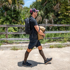 A man in athletic clothes is walking outdoors, carrying a tan Sand Tombstones workout sandbag and wearing a black backpack and cap.