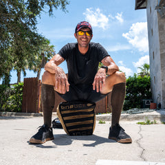 Wearing sunglasses and a smile, a man in sportswear squats outdoors on a Sand Tombstones loadable sandbag on a sunny day.